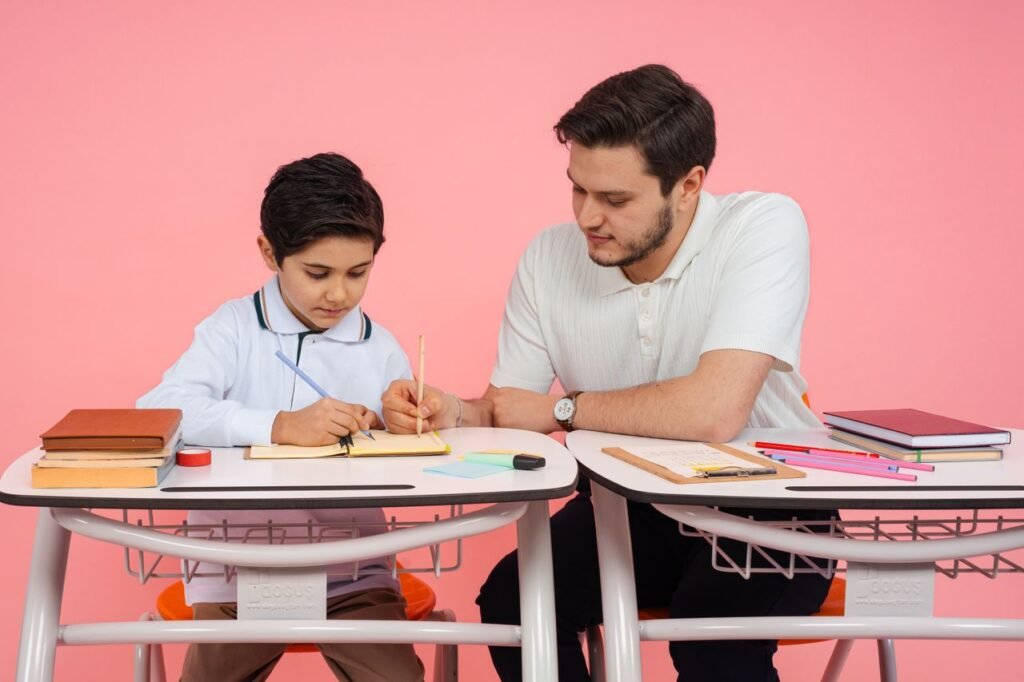 A teacher helps a student with schoolwork at a desk against a pink background.